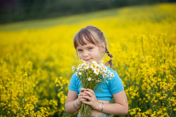 Cute little blonde girl blue dress in boho style in rapeseed field holding in hand bouquet of chamomiles in summer day.