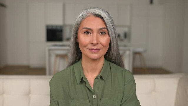 Headshot of charming gray-haired 50s Asian woman looking at the camera and starting break into a smile, video portrait of attractive positive mature lady in home interior