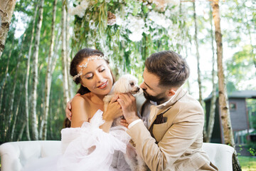 Luxury bride and groom are playing with a fluffy white dog, sitting on a sofa in the forest. 