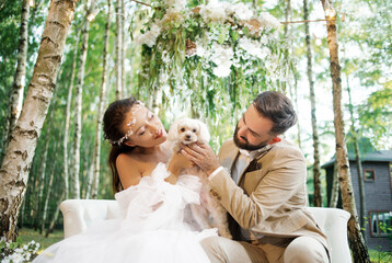 The bride and groom are playing with a fluffy white dog, sitting on a sofa in the forest. 