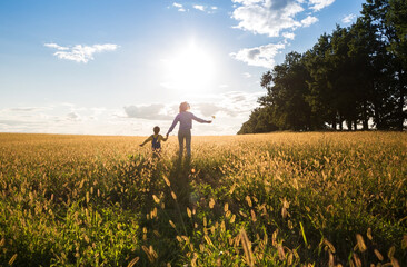 unrecognizable child and woman, mother and son, run across a yellow meadow towards the sun. Happy...