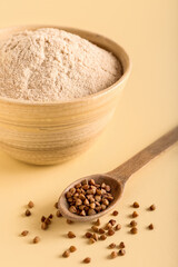 Bowl of flour and spoon with buckwheat grains on beige background