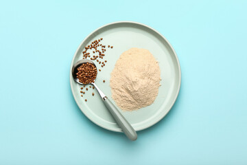 Plate of flour and spoon with buckwheat grains on blue background