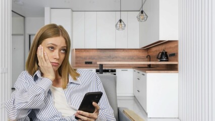 Young bored woman watching smartphone sitting in chair at home. Caucasian girl resting and using...