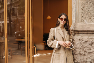Obraz premium Stylish young caucasian woman in sunglasses holding glass of drink standing near cafe during daytime. Brunette wears long beige jacket. Lifestyle, different emotions, leisure concept.