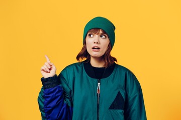 a laughing, happy woman in a green jacket and a hat in the style of the 90s stands on a yellow background, smiling happily and pointing to an empty space for inserting an advertising layout