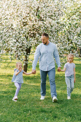 Fototapeta premium Happy family. A father and two daughters walk through the park in spring in a blooming garden, holding hands