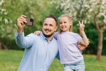 Dad and his six-year-old daughter take selfies on the phone, smiling and having fun on camera in the park in summer or spring