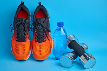 Orange sneakers, dumbbells and plastic drinking water bottle on a blue background. Fitness and weight loss equipment