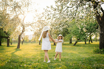 Naklejka na ściany i meble A cute mom in a hat and a white dress is walking through the spring garden with her cute daughter. Mom and baby are having fun in spring flowers. Family together outdoors.