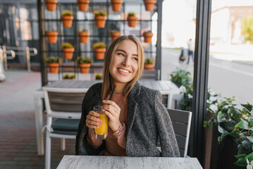 A happy young woman is sitting at a table in a summer cafe and holding a glass of mango juice in her hand