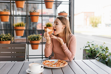 A happy young woman is eating a slice of pizza in a summer cafe at a table