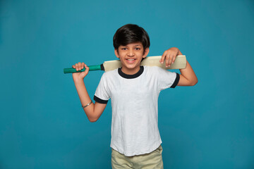 Portrait of smiling boy standing with cricket bat on shoulder against blue background