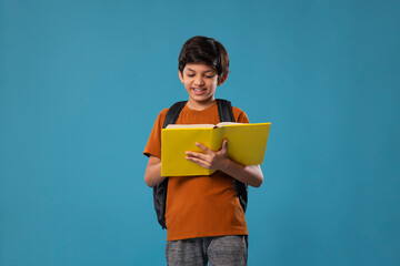 Boy reading book while standing with backpack against blue background