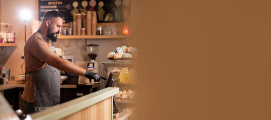 Bearded waiter in apron at counter with cashbox working at bar or coffee shop, male barista at counter in cafe store checking client's order, small business, people and service concept