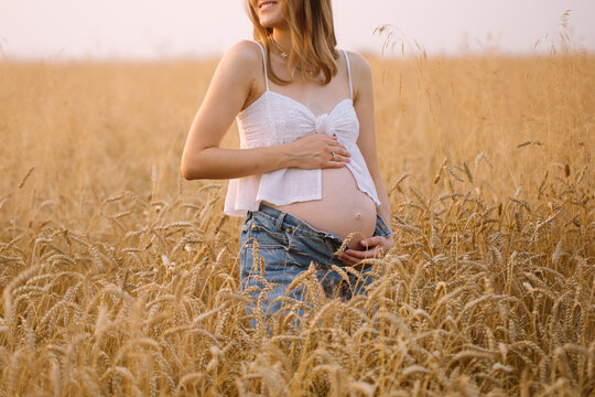 A pregnant woman with blond hair in a white top and denim shorts is spinning in a wheat field. The girl strokes her pregnant belly.