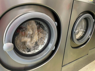 Dirty clothes spinning inside the main doors of two stainless steel industrial washing machines.