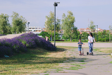 Naklejka premium Happy family lifestyle and holiday concept. Mother, little boy riding scooters, walking in old city, street. Laughing on a summer sunny day. Having fun