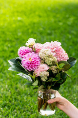 Hand holding a glass jar with bouquet of garden flowers on a green grass background