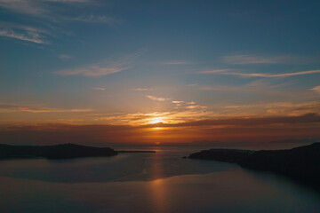Landscape orientation image a beautiful vibrant and colourful sunset in Santorini. Aegean sea in foreground, sun setting near Oia in the distance. - obrazy, fototapety, plakaty