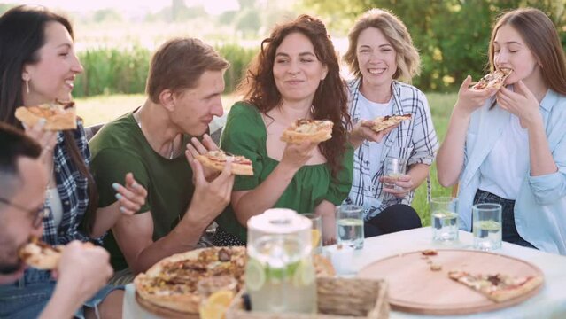 Group of friends are relaxing together outdoor. Young people are sitting at the table, having fun talking and eating pizza.