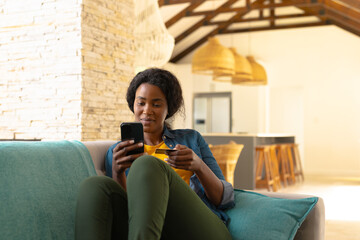 African american young woman holding credit card while online shopping through smart phone on sofa