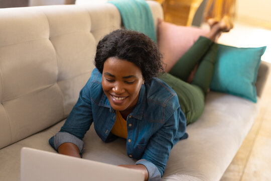 Smiling young african american woman using laptop while lying on sofa at home, copy space