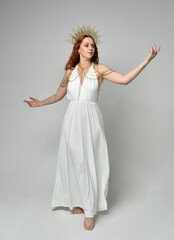 Full length portrait of beautiful red head woman wearing long flowing fantasy toga gown with golden halo crown jewellery, standing pose   isolated on a white studio background.
