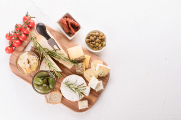Selection of cheeses served with variety of antipasti - olives, baby and sun dried tomatoes, pickled artichokes, capers and crackers at white table background with copy space. Cheese plate