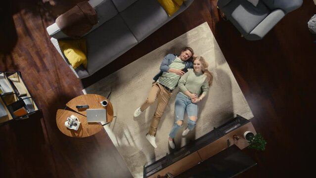 Top View Apartment: Happy Couple Relaxing on a Carpet in the Living Room, Using Smartphone. Girlfriend and Boyfriend Enjoy being Together, Online Shopping, e-business, Browsing Social Media. Zoom Out