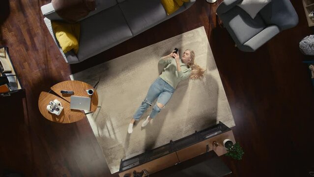 Apartment: Young Woman Lying on a Living Room Floor, Using Smartphone. Girl Relaxes at Home. Freelancer Does Remote Work, E-business, Online Shopping, Social Media Browsing. Top View Static Shot