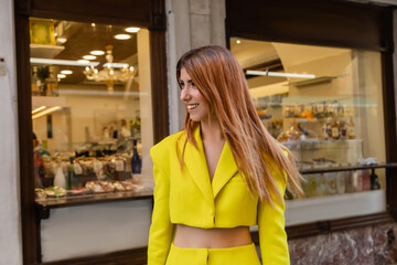 stylish redhead woman in yellow clothes smiling near building with shops in Venice.