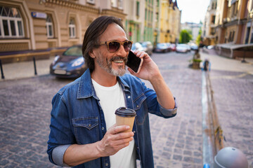 Happy mature grey bearded man talking on the phone, having coffee in paper cup or lunch break during work or travel time standing outdoors in urban old city background wearing jeans shirt