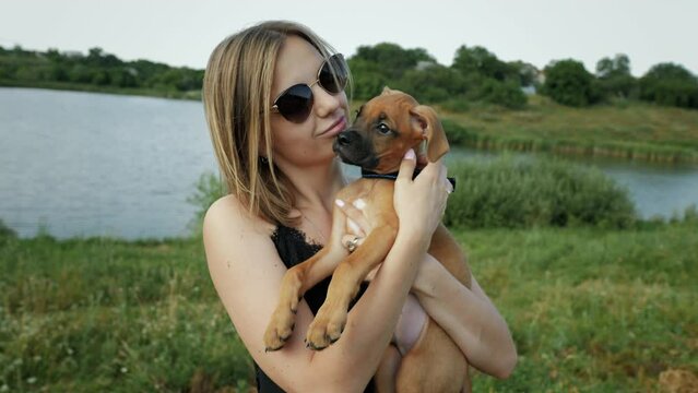 Woman holds on arms and hugs a small german boxer puppy in nature near the lake