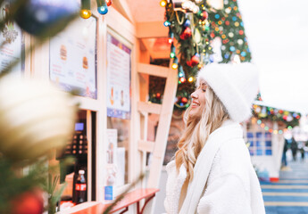 Young happy woman with curly hair in white knitted hat on shopping at the Christmas fair market in winter street decorated with lights