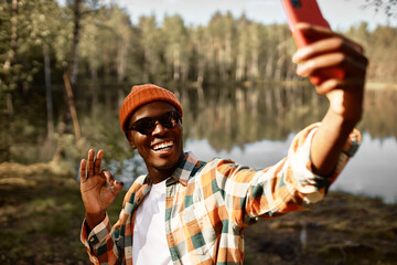 Outdoor image of black man showing ok sign during video chat with his family standing against lake...