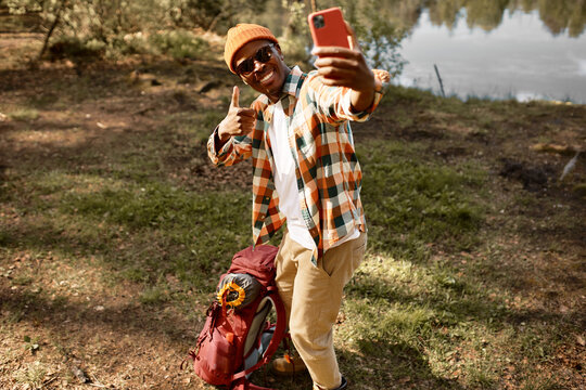 Image of cheerful african american man wearing hat making selfie on phone in wild nature near river showing thumbs up gesture, smiling video chatting with his friends or family, sharing good vibes - Powered by Adobe