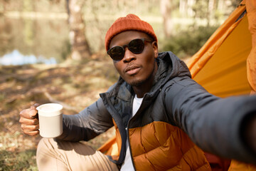 African american man hiker in spring coat and sunglasses taking selfie on smartphone sitting near his orange tent by river in forest, holding white cup of tea. Traveling, adventures, technology