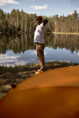 Fototapeta na wymiar Vertical side view image of happy relaxed african american man hiker stretching hands in morning standing near lake with sky and forest reflection, after spending night in tent. Camping concept