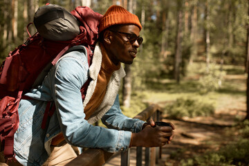 Fototapeta premium Happy african american guy in sunglasses and denim coat having rest leaning on wooden bridge in national park, admiring beauty of wild nature,with backpack on shoulders, holding thermos mug of tea