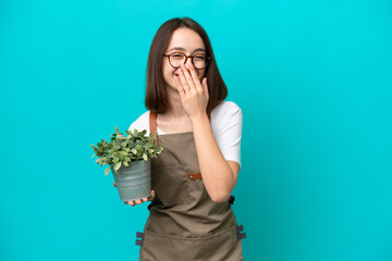 Gardener Ukrainian woman holding a plant isolated on blue background happy and smiling covering mouth with hand