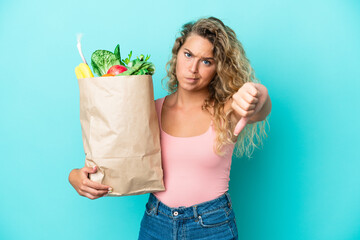 Girl with curly hair holding a grocery shopping bag isolated on green background showing thumb down with negative expression