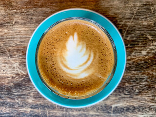A cup of coffee latte On a wooden table. A mug of flat white coffee on a wooden background. Coffee art. Heart flower shape latte art. Copy space. Glass on turquoise blue saucer