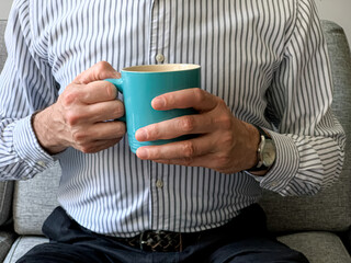 Young businessman freelancer is drinking morning coffee at home blue ceramic mug sitting on sofa. Young businessman freelancer is drinking morning coffee at home blue ceramic mug sitting on sofa