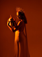 Close up portrait of beautiful red-haired woman wearing long flowing fantasy toga gown with golden halo crown jewellery,  creative hand gestures on a dark moody background with glowing orange lighting