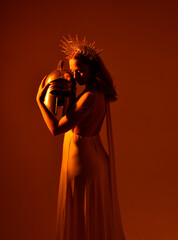 Close up portrait of beautiful red-haired woman wearing long flowing fantasy toga gown with golden halo crown jewellery,  creative hand gestures on a dark moody background with glowing orange lighting