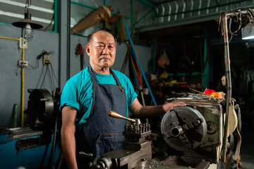 Fototapeta na wymiar A senior Asian craftsman, standing closely and carefully controlling a lathe, inside a small factory of his family business..