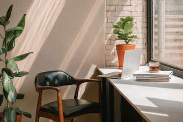 Inspiring workplace near window. Laptop, books, coffee cup, leather chair, plants. Sun rays on wall. Home office.