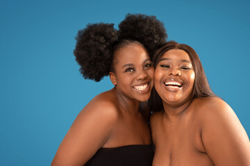 Two beautiful happy women with amazing toothy smiles posing together on blue studio background, looking at the camera. Real people emotions.