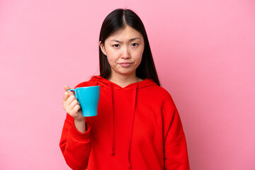 Young Chinese woman holding cup of coffee isolated on pink background with sad expression